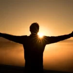 Man with arms open at sunset with the sunset beaming past shoulder