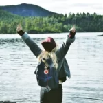 A woman standing by a lake with their arms in the air, trees in the background, celebrating an achievement