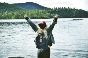 A woman standing by a lake with their arms in the air, trees in the background, celebrating an achievement