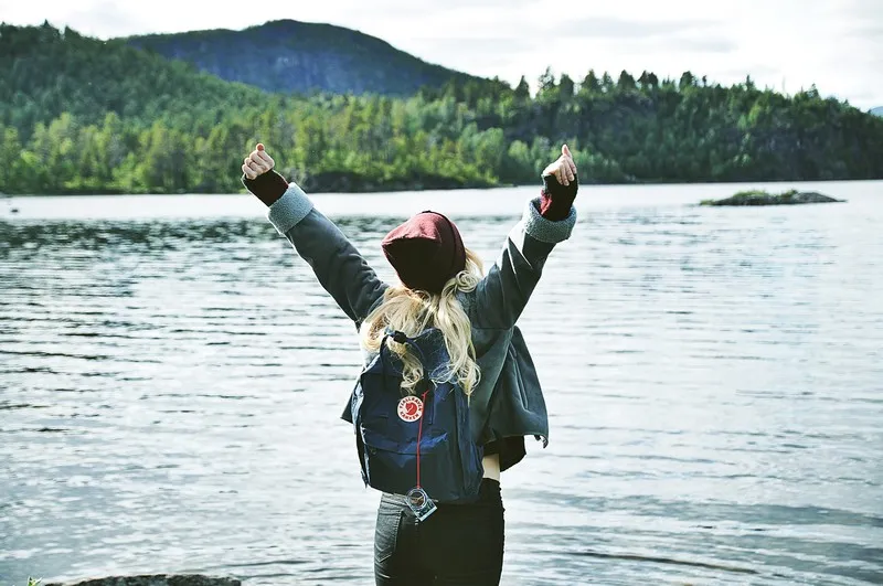 A woman standing by a lake with their arms in the air, trees in the background, celebrating an achievement