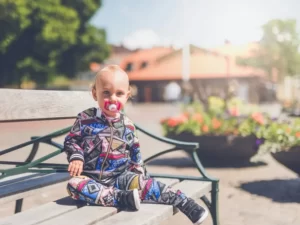 A toddler sat on a public bench on a sunny day with a pacifier / dummy in its mouth