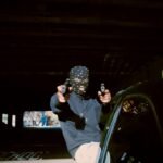 A man wearing a balaclava at night and pointing two guns akimbo at the camera