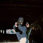 A man wearing a balaclava at night and pointing two guns akimbo at the camera