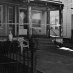 a black and white crime scene showing the front facade of a house cordoned off with police tape - neoliberalism