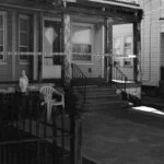 a black and white crime scene showing the front facade of a house cordoned off with police tape - neoliberalism