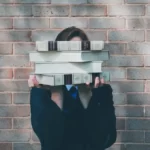 A student stood in front of a wall hiding behind a pile of books