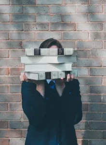 A student stood in front of a wall hiding behind a pile of books