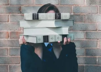 A student stood in front of a wall hiding behind a pile of books