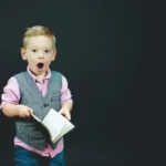 A smart looking young boy holding a book and looking shocked