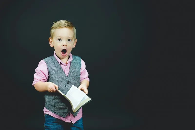 A smart looking young boy holding a book and looking shocked