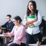 A student in a education classroom standing up. Other students are sat down.