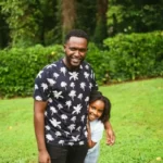 A black man and his black daughter standing in a park.