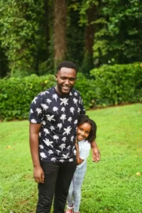 A black man and his black daughter standing in a park.