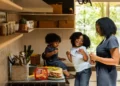 A black mother and her two children in the kitchen. one child is sat on the counter top. neoliberal family