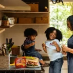 A black mother and her two children in the kitchen. one child is sat on the counter top. neoliberal family