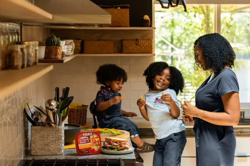 A black mother and her two children in the kitchen. one child is sat on the counter top. neoliberal family