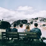 five friends sat on a wooden bench viewed from behind and looking out over towards a city - social capital