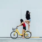 two women riding a bike - one stood on the back wheel - feminism