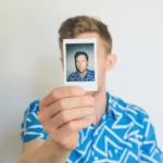 a man in a blue shirt holding up a polaroid of himself in front of his face - identity