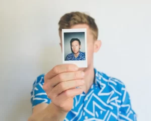 a man in a blue shirt holding up a polaroid of himself in front of his face - identity