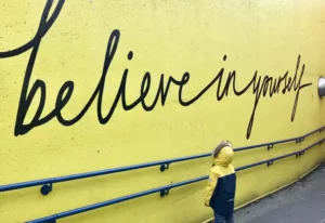 a small boy looking at a yellow wall with the words 'believe in yourself' - self-concept