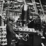 a black and white shot of a man wearing a flat cap operating a historical machine