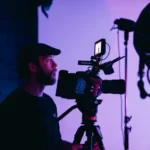 A man behind a TV camera in front of a dimmed purple background.