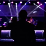 A music producer stood at a mixing desk against a purple lighted background stage lights