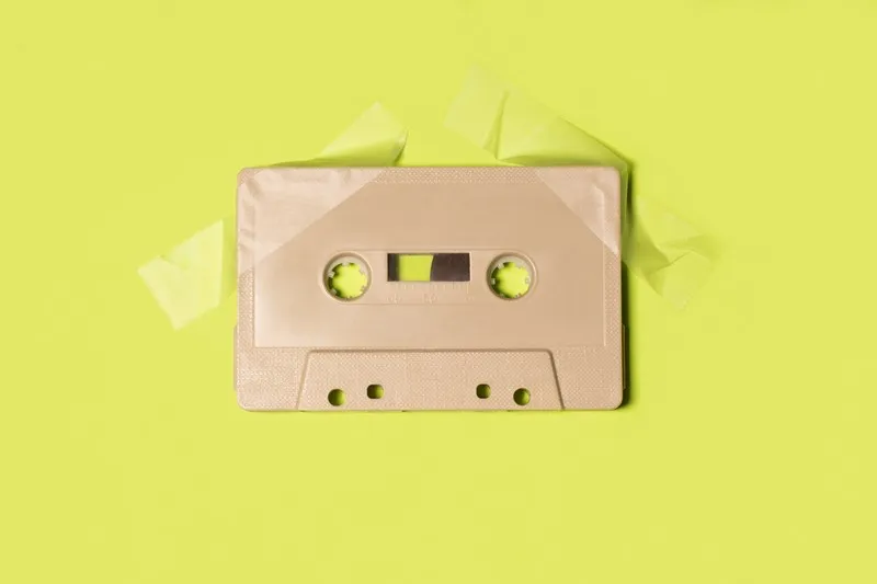 A beige cassette tape attached to a yellowy-green background with sticky tape