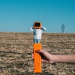 a person holding an orange TV remote and pointing it at a man with an orange TV on his head