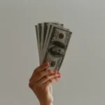 A hand offering a wad of dollar bills against a grey background