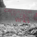black and white shot of a grim wall. has red writing stating 'capitalism is the virus'. neoliberalism.