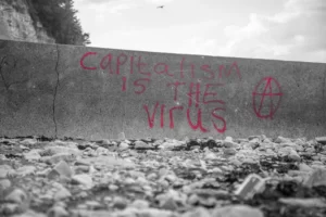black and white shot of a grim wall. has red writing stating 'capitalism is the virus'. neoliberalism.