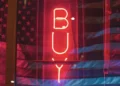 A red neon sign in the darkness which reads 'buy' - privatisation for profit