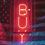 A red neon sign in the darkness which reads 'buy' - privatisation for profit