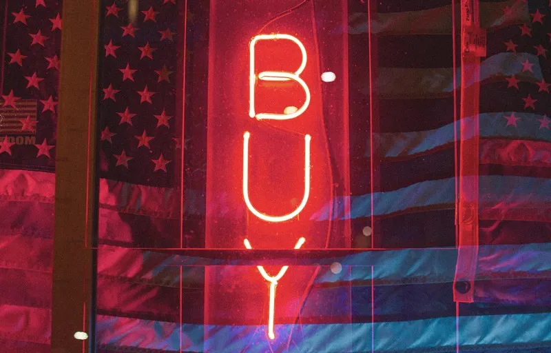 A red neon sign in the darkness which reads 'buy' - privatisation for profit