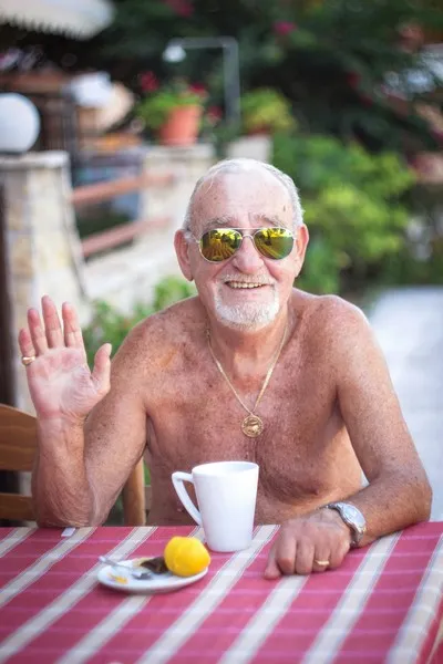 A shirtless older man waving with a cup of tea