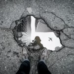 black and white shot of a pothole filled with water and reflecting an aeroplane in the sky