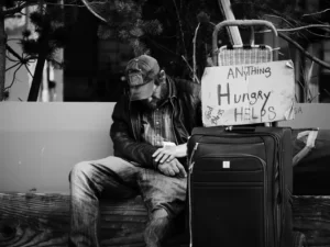 an oppressed homeless man sat on a bench with a suitcase and a sign saying 'hungry, anything helps'