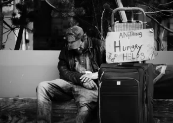 an oppressed homeless man sat on a bench with a suitcase and a sign saying 'hungry, anything helps'