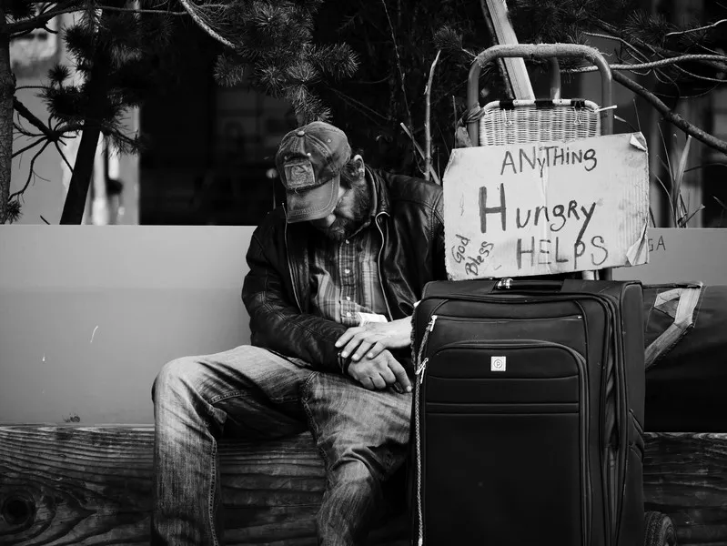 an oppressed homeless man sat on a bench with a suitcase and a sign saying 'hungry, anything helps'