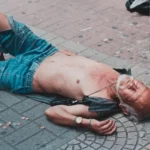 A homeless man laid out on a cobbled floor with no shirt on experiencing accumulation by dispossession