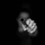 a blurred person aiming the barrel of a gun directly towards the camera against a black background