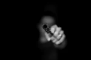 a blurred person aiming the barrel of a gun directly towards the camera against a black background