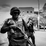 A person armed with a shotgun and wearing flak jacket and bullet belt