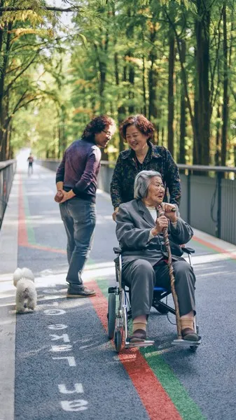 An aging woman in a wheelchair on a sunny day. She is surround by trees and shade.