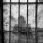 the silhouette of a person behind bars in a jail cell