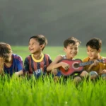four young boys sat in a field smiling with one playing a guitar