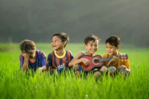 four young boys sat in a field smiling with one playing a guitar