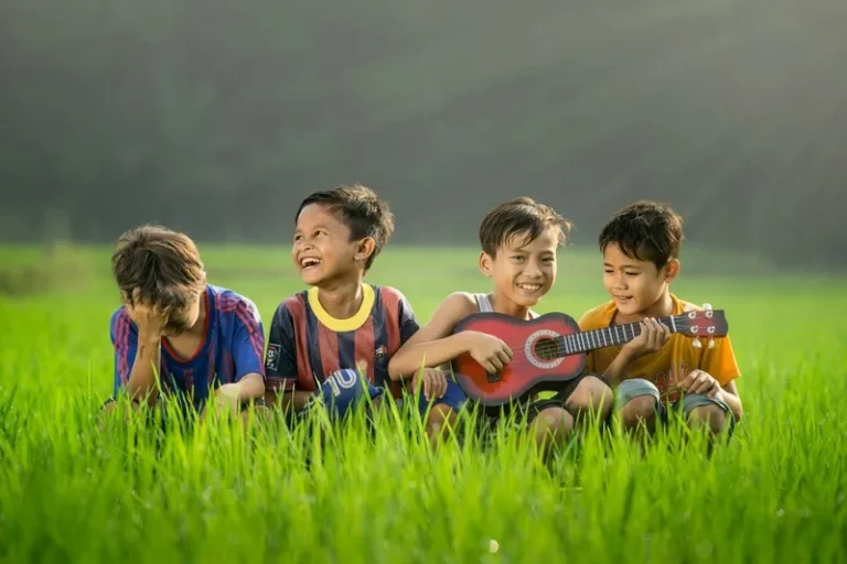 four young boys sat in a field smiling with one playing a guitar